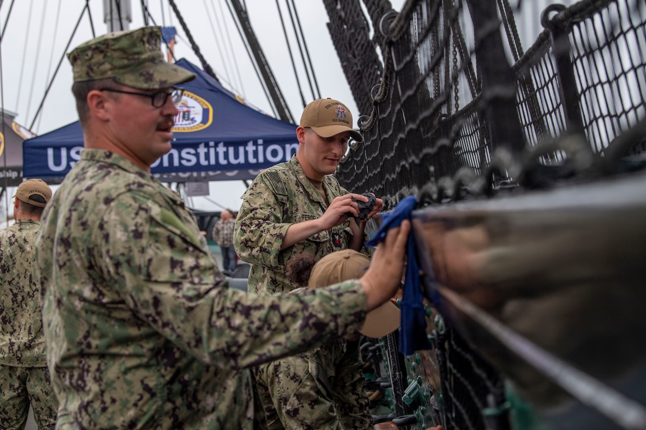 Two sailors polish the railing of a ship while on deck.