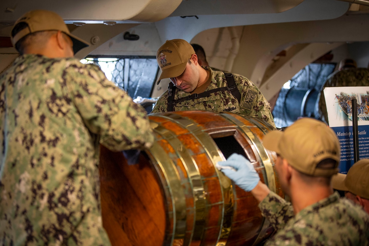 A group of sailors polish a wooden barrel on a ship.
