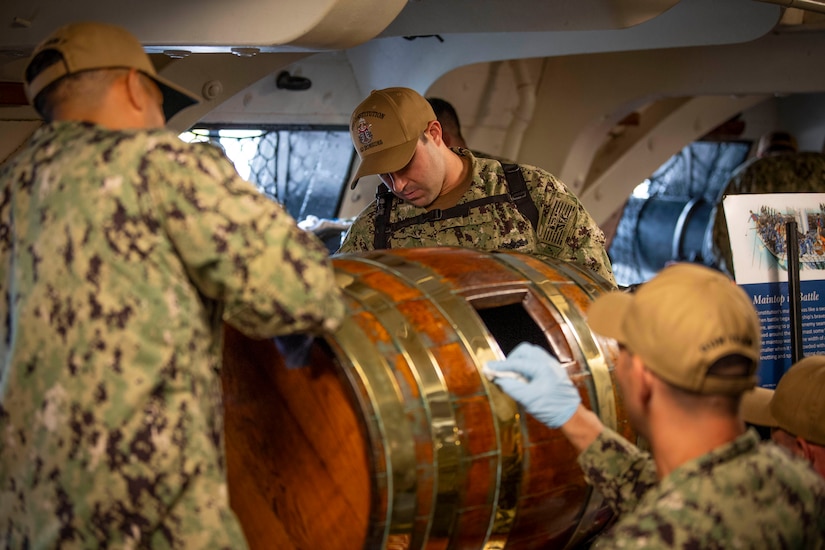 A group of sailors polish a wooden barrel on a ship.