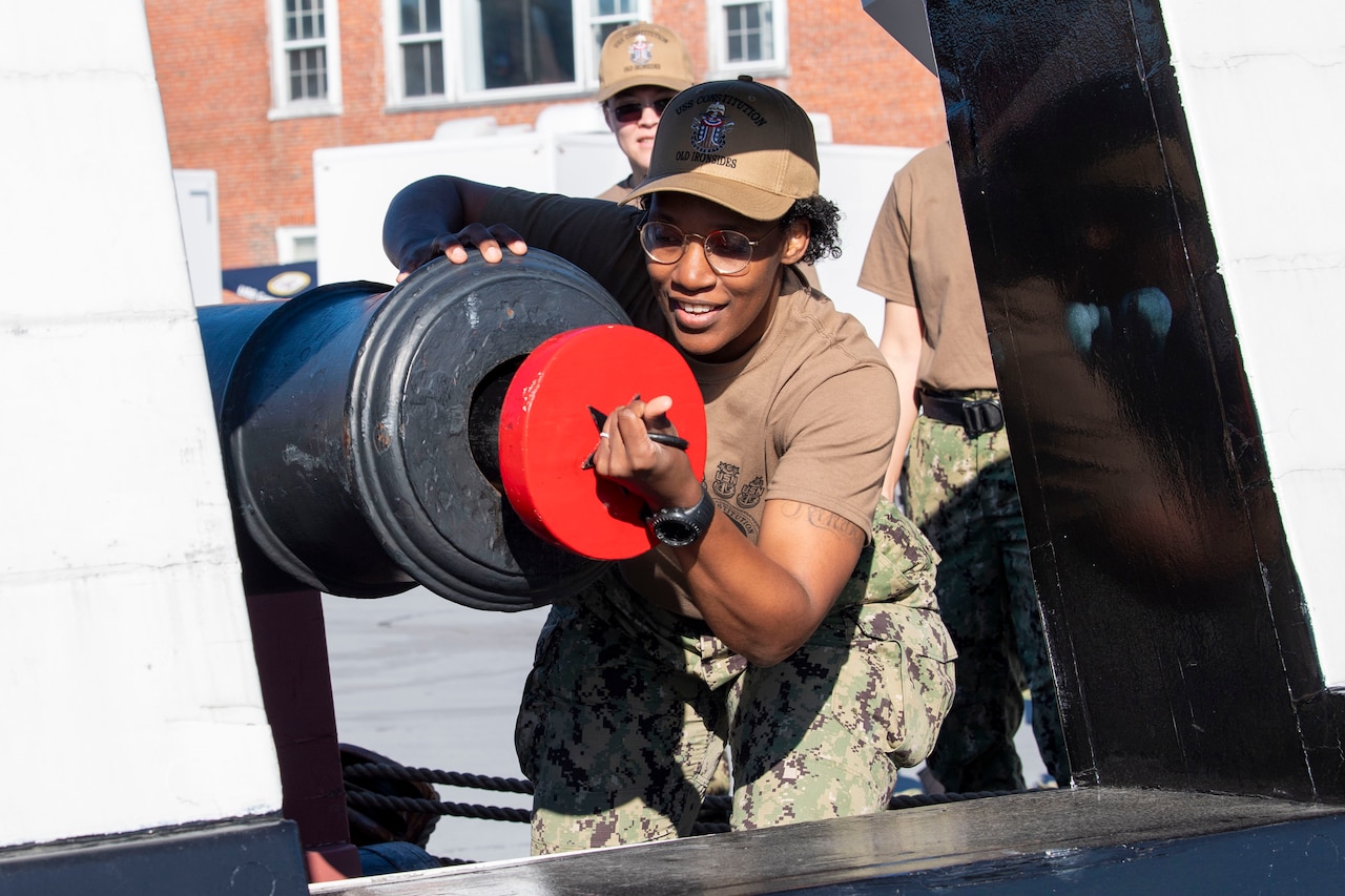 A sailor cleans a canon with an orange brush.