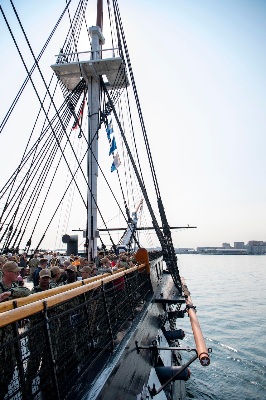 An old ship sails into the bay while people on deck watch.