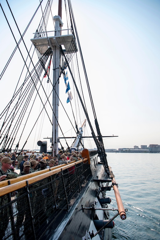 An old ship sails into the bay while people on deck watch.