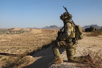 A soldier knees atop a hill over looking a desert valley during the day.