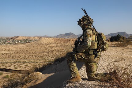 A soldier knees atop a hill over looking a desert valley during the day.