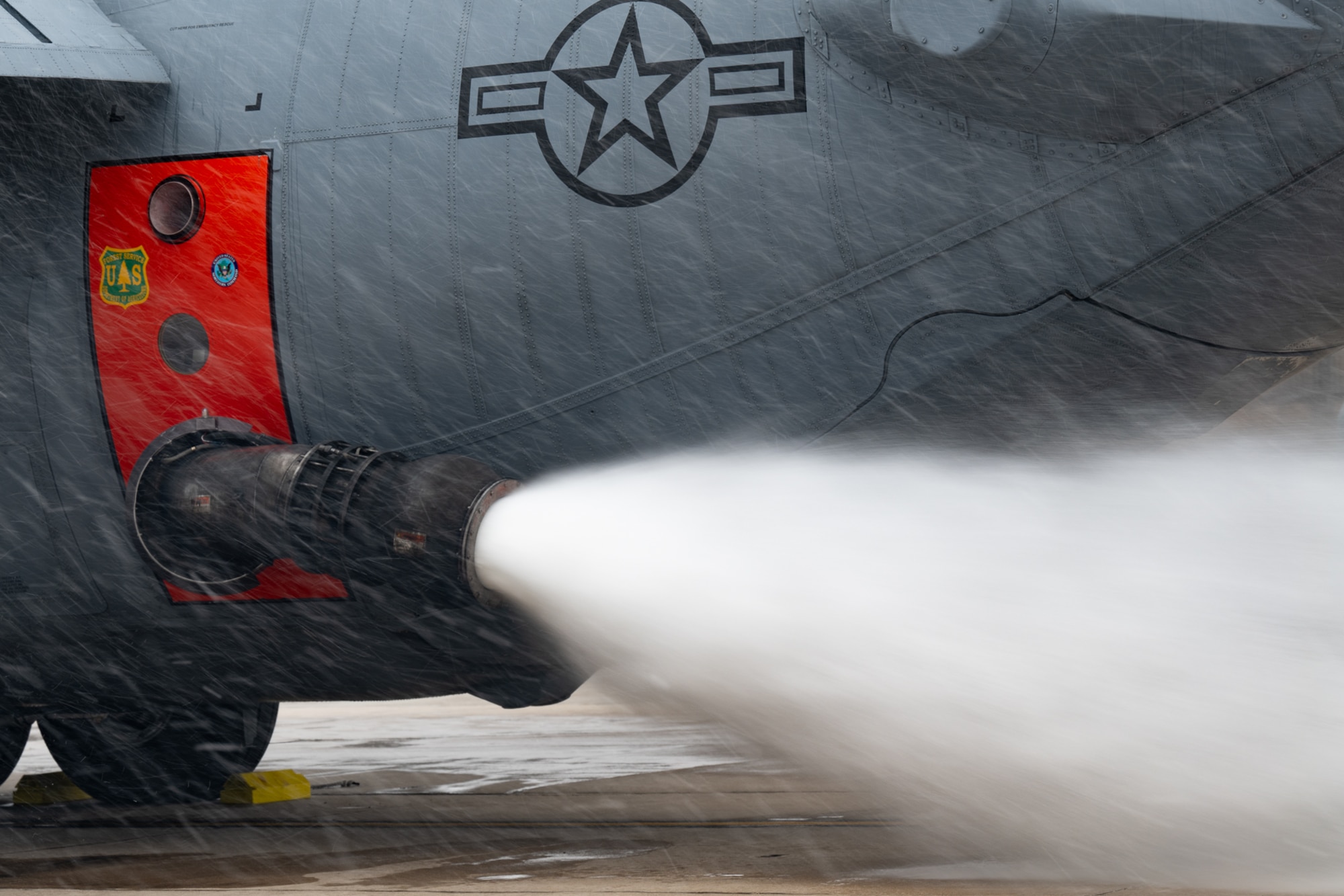 A nozzle protruding from the side of a military aircraft emits a burst of water onto the concrete.