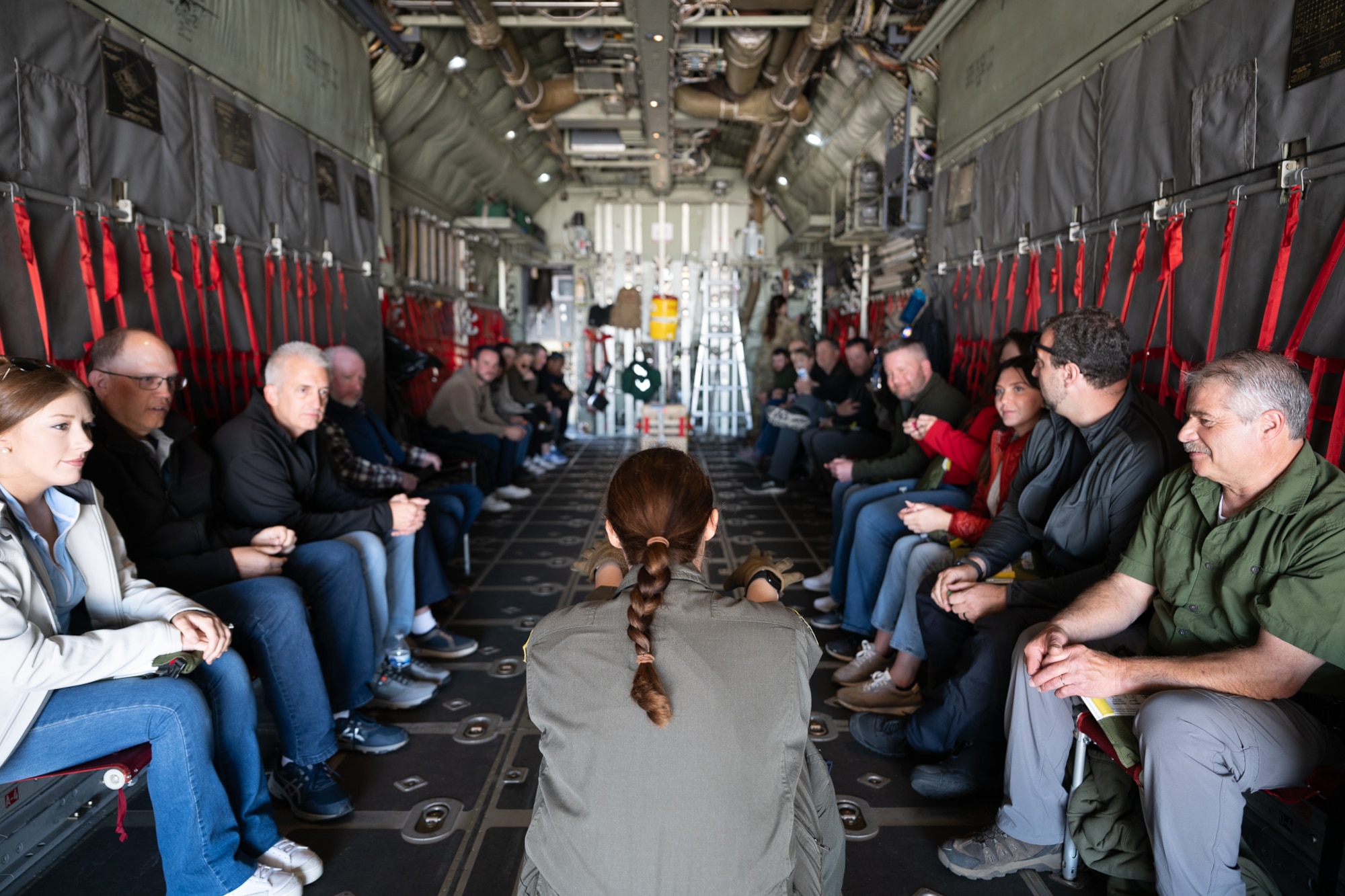 Civilians seated in chairs in a military aircraft listening to a woman in a flight suit.