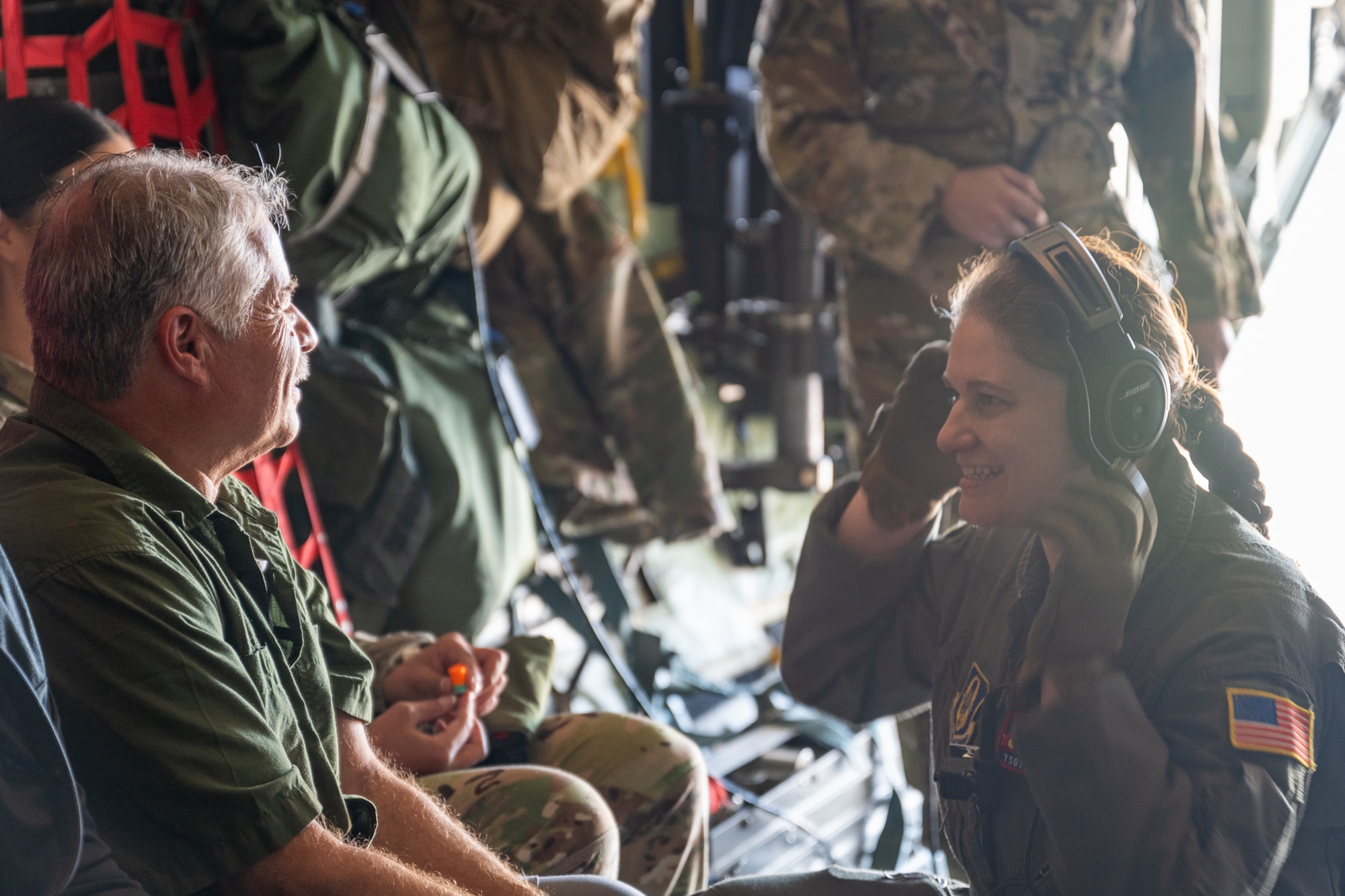 A man in civilian clothes speaks to a woman wearing a flight suit and a headset with a brightly lit background in an aircraft.