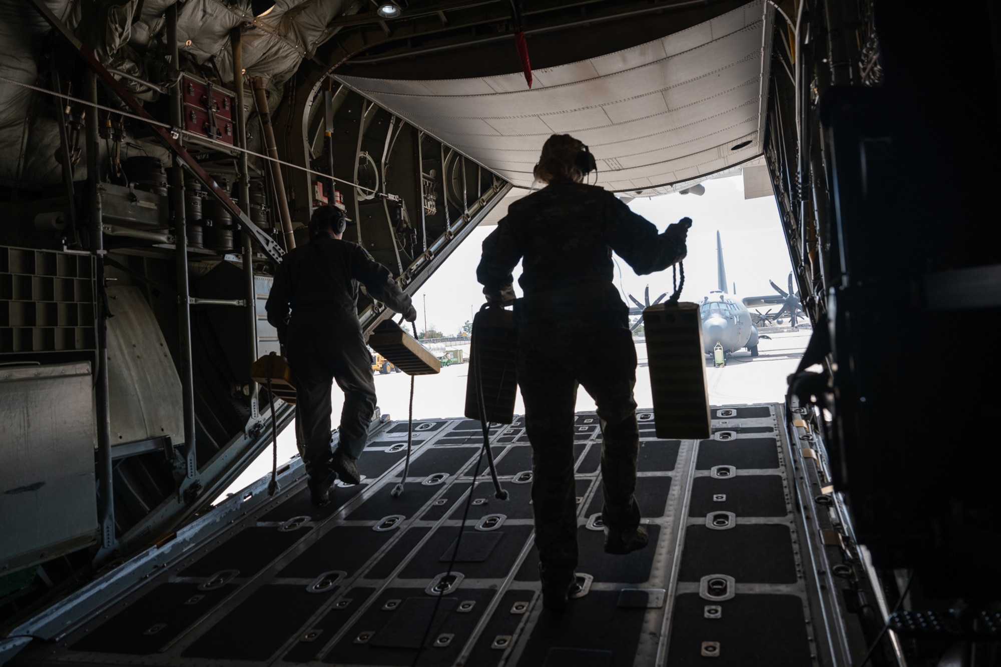 Silhouettes of two people in flight suits walking towards the rear of a cargo bay holding parking chocks with an aircraft in the background.