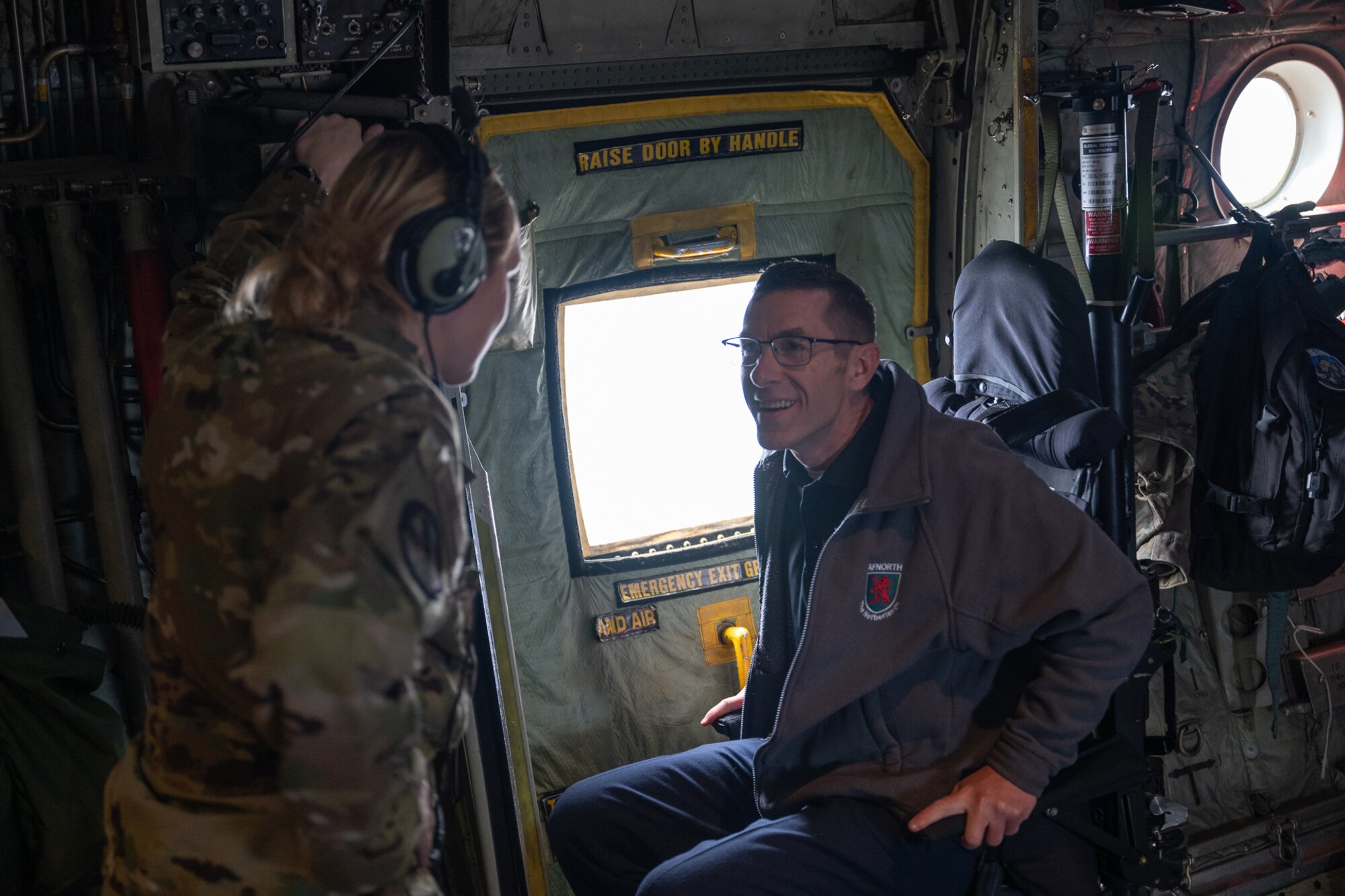 A woman in military uniform speaks to a man seated in a chair on the back of an aircraft.