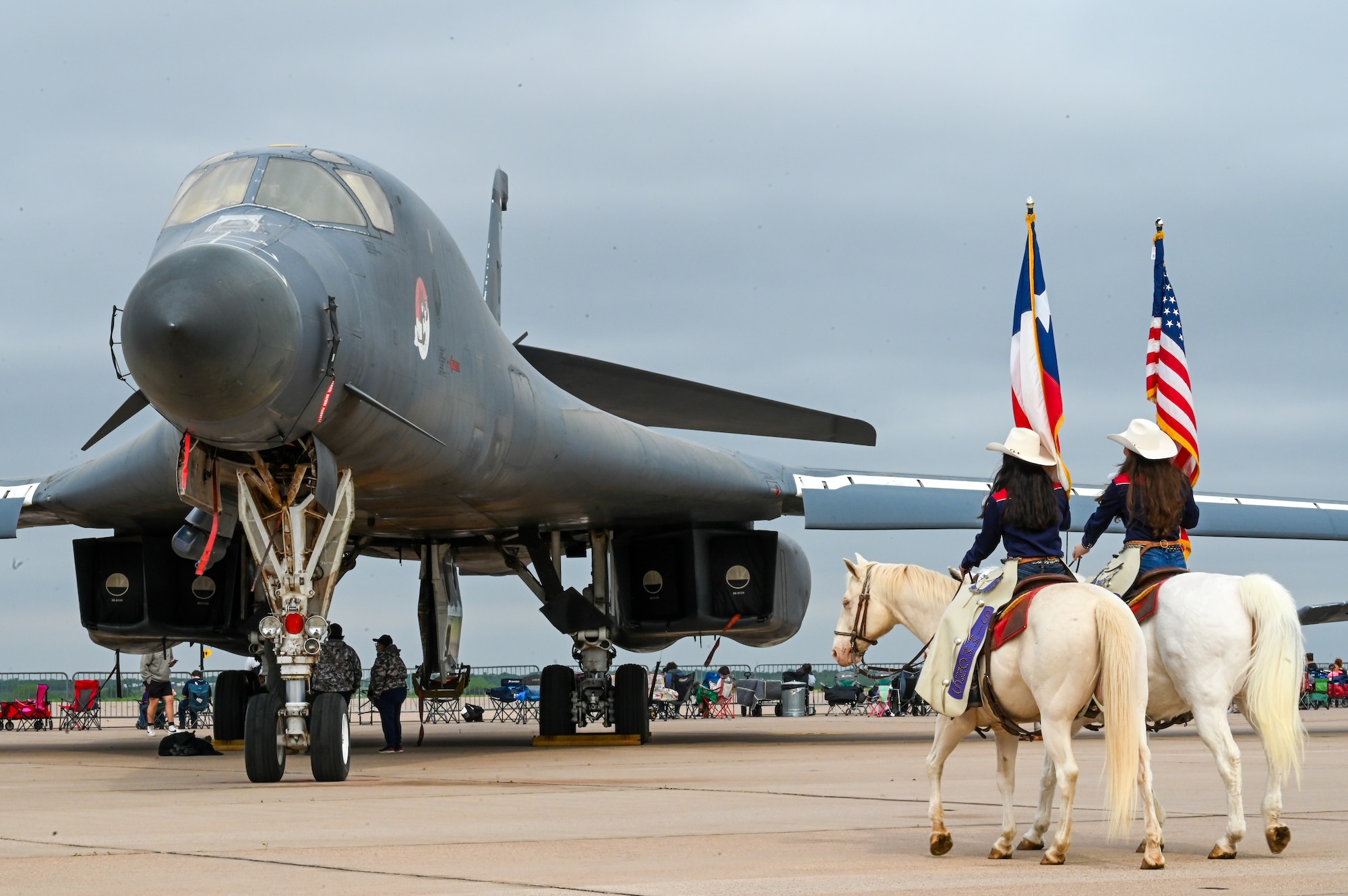 two white horses with riders holding the Texas and American flags walk in front of a B-1 on the flightline