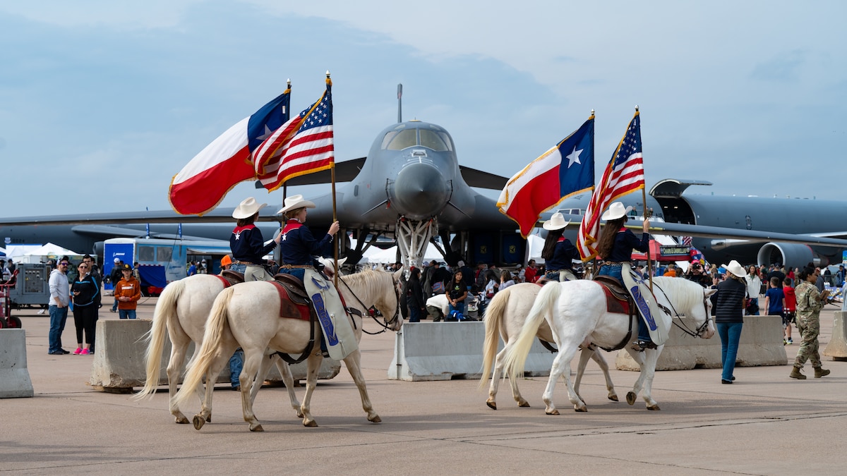 Four white horses with female riders holding the Texas and American flags walk in front of a B-1 Lancer