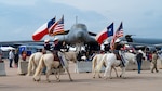 Four white horses with female riders holding the Texas and American flags walk in front of a B-1 Lancer