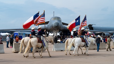 Four white horses with female riders holding the Texas and American flags walk in front of a B-1 Lancer