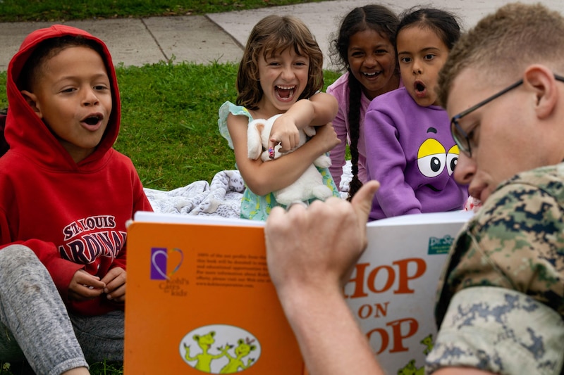 A Marine sitting in grass holds up a book while four children laugh and one points.