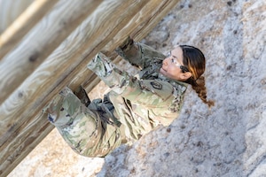 A soldier climbs up a wooden obstacle while wearing safety goggles.
