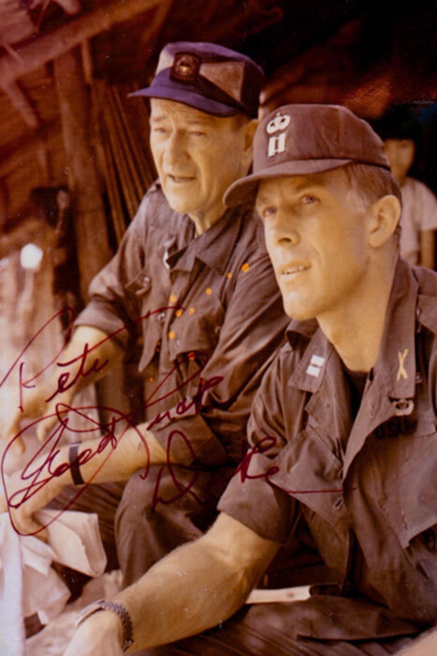 A soldier in a military uniform sits next to another man wearing a hat and a button-up shirt.