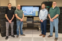 Four men pose for a photo in front of a large television.