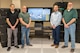 Four men pose for a photo in front of a large television.