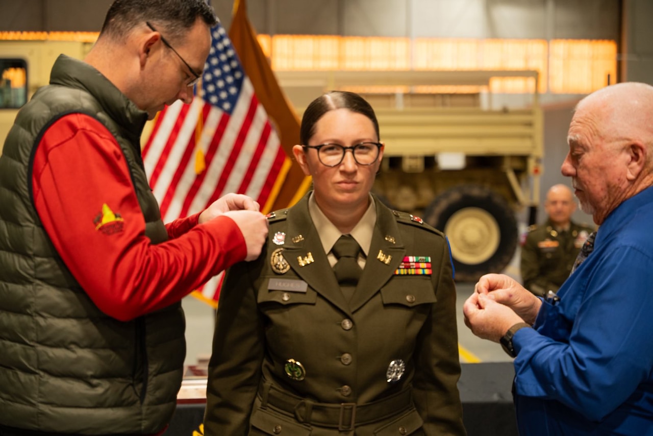 Two people pin a soldier wearing formal attire indoors in front of flags.