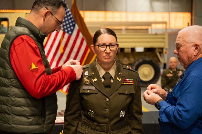Two people pin a soldier wearing formal attire indoors in front of flags.