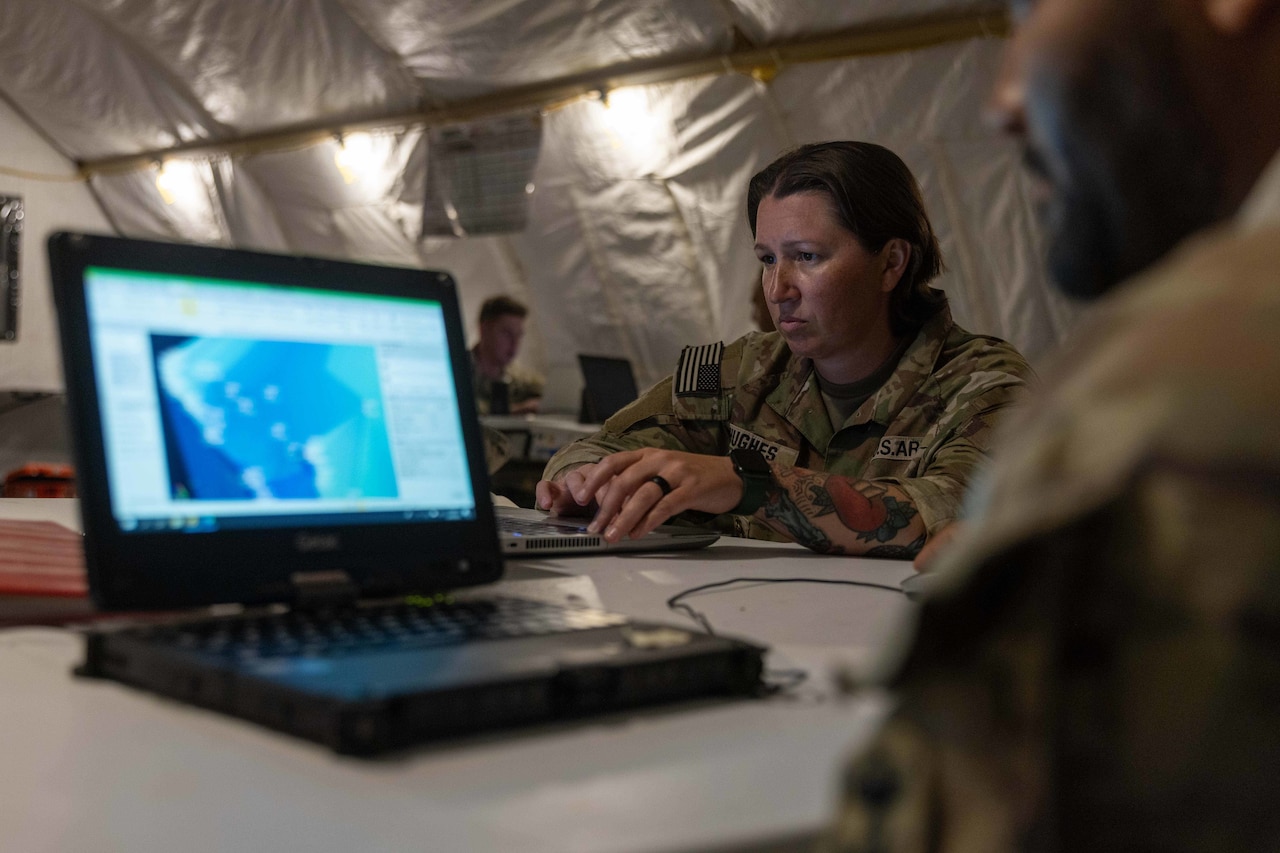 Two soldiers sit in front of laptops and work inside a tent.