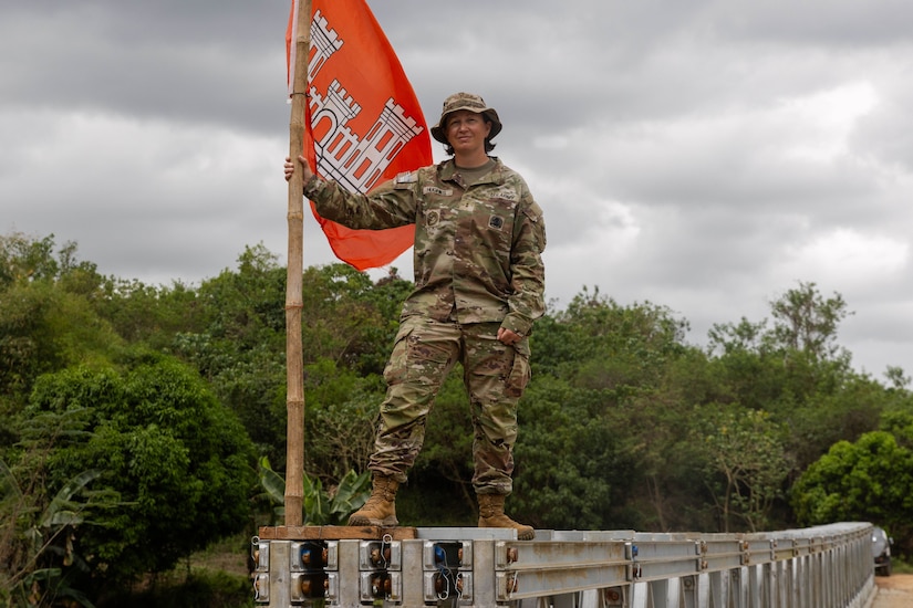An Army engineer holds the army's engineering flag while standing on metal on a field next to trees in an overcast day.