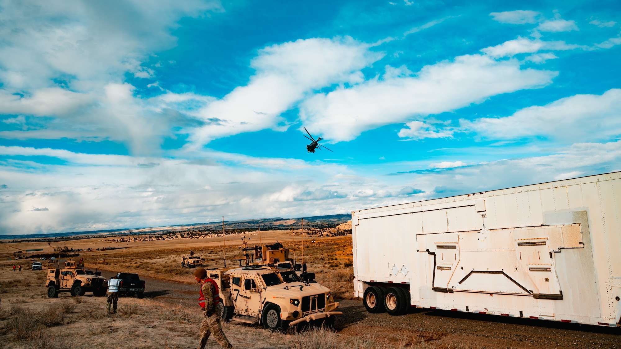 Military vehicles and military personnel are on a road while a helicopter flies in the background