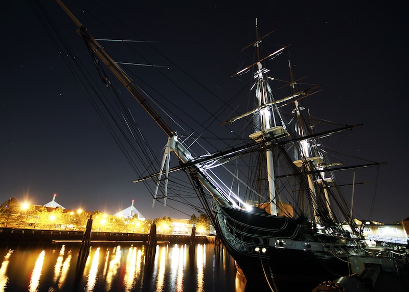 An old ship is moored to a pier at night while lights shine from the left.