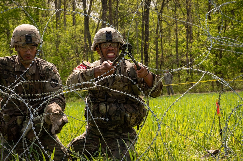 Two Soldiers are next to barbed wire. One Soldier on the right is cutting it with wire cutters while the other uses special gloves to hold it in place.