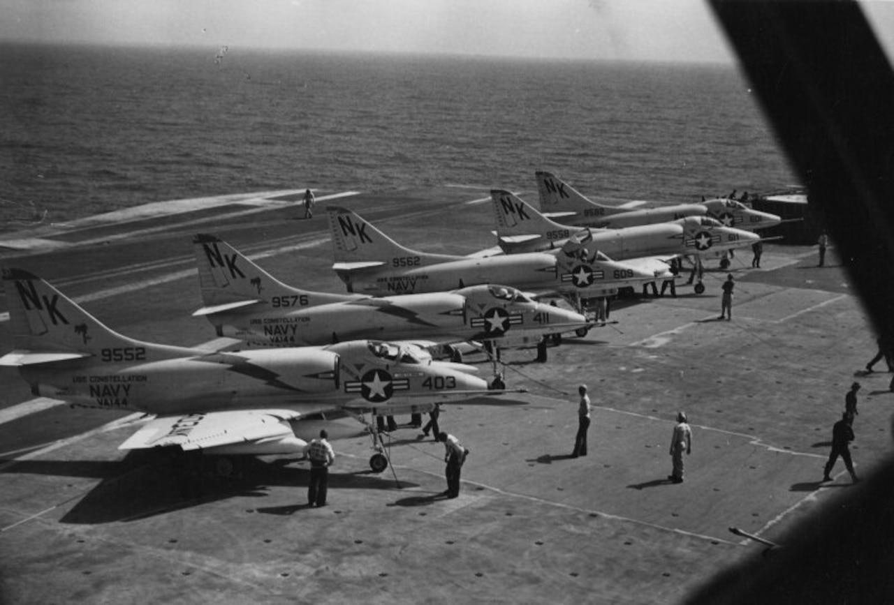Five aircraft sit on the deck of an aircraft carrier as people work around them.