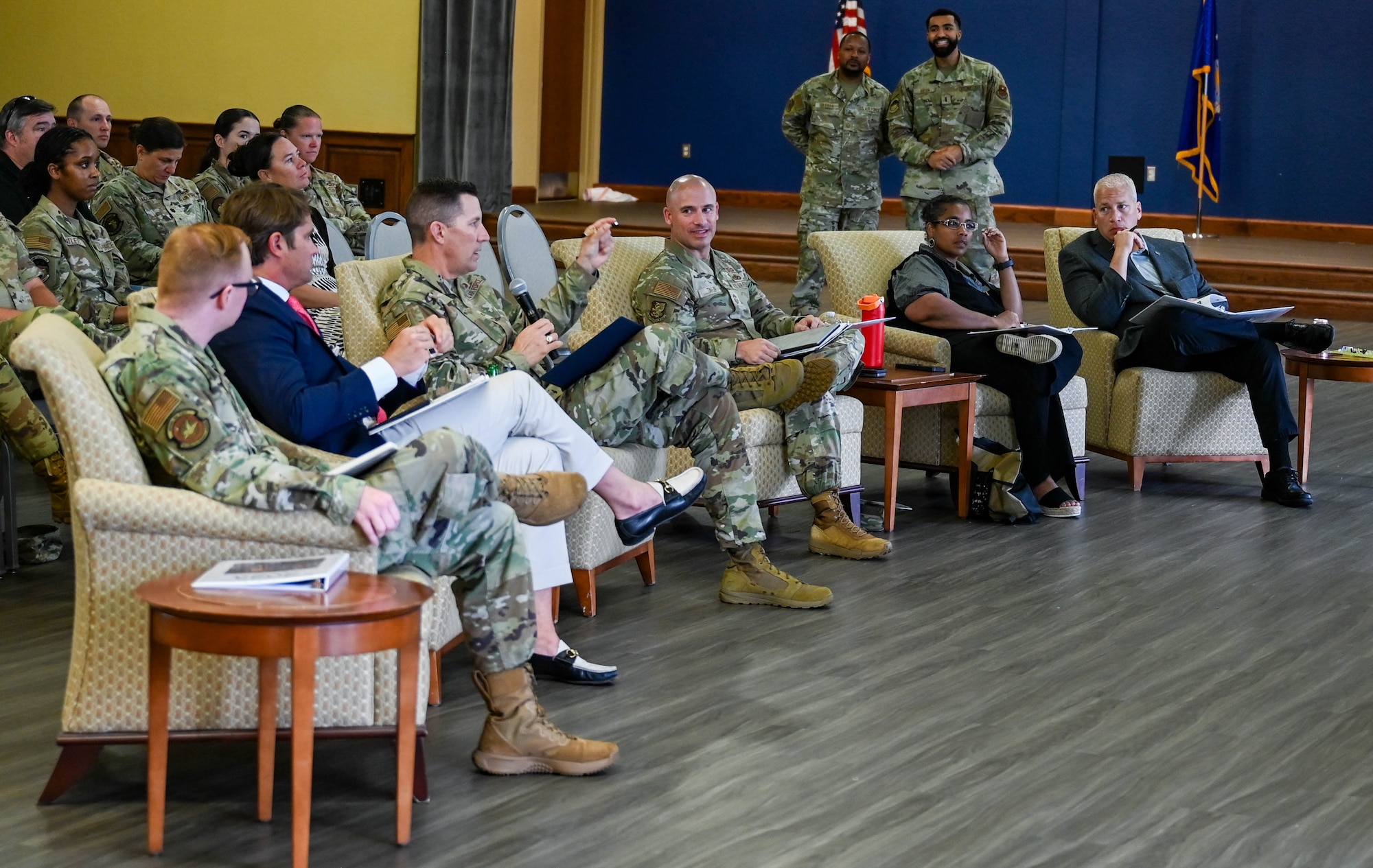 Six people sit in chairs during the Dragon Tank competition panel, with a uniformed male member, holds a microphone while speaking to presenters.