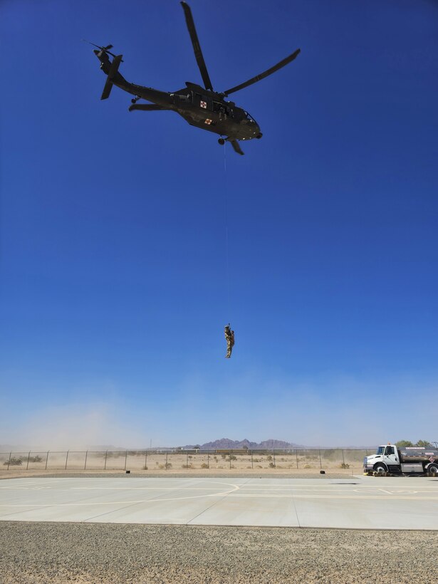A soldier hangs from a hovering helicopter under a blue ski close to desert terrain.