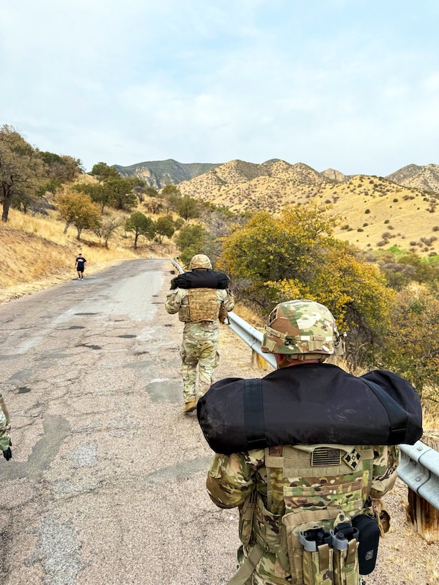 Two soldiers carry gear and extra weight up a mountain road.