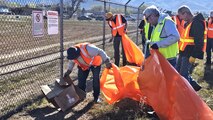 Volunteers, some holding trash bags, pick up trash along a fence line.