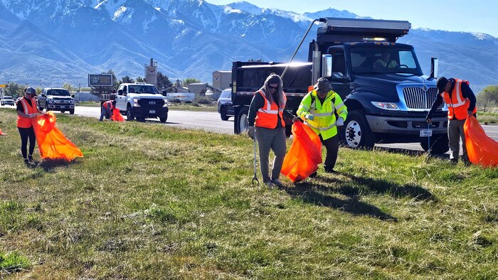 Volunteers, some holding trash bags, pick up trash along a fence line.