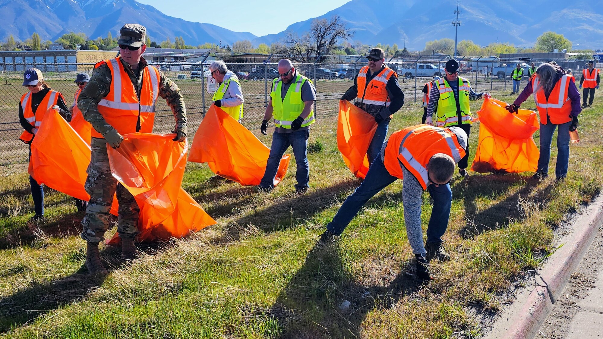 Volunteers, some holding trash bags, pick up trash along a fence line.