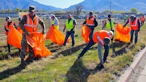 Volunteers, some holding trash bags, pick up trash along a fence line.