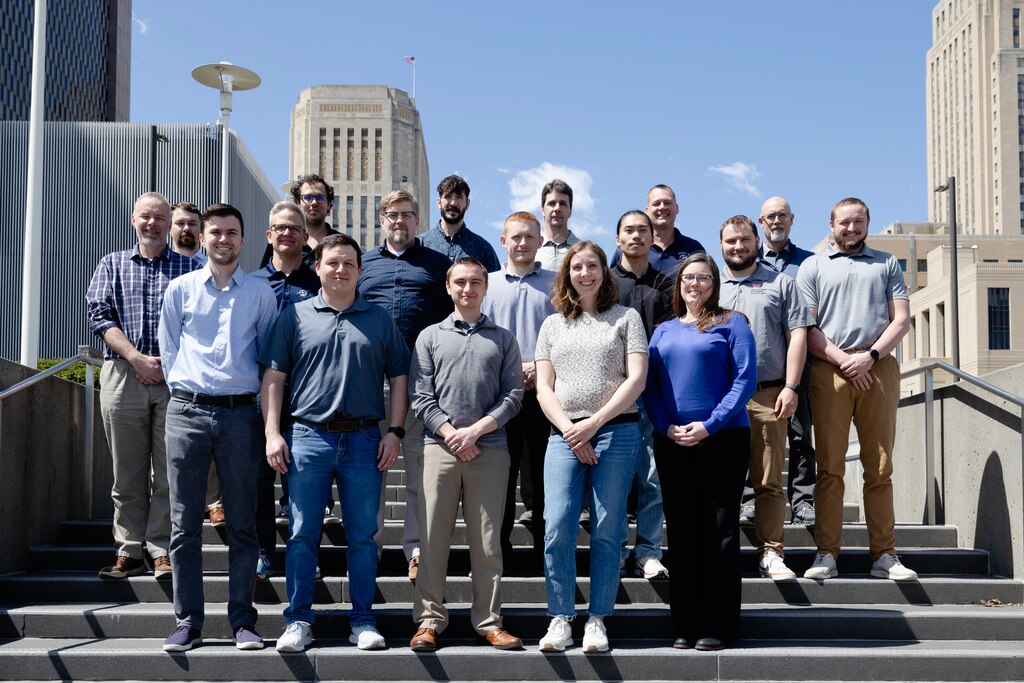 A group of people stand staggered on steps outside of a building with a blue sky in the background.
