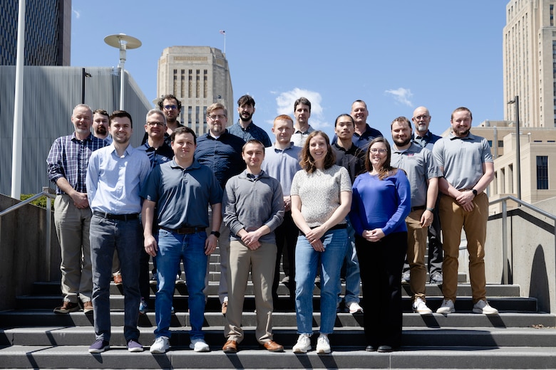 A group of people stand staggered on steps outside of a building with a blue sky in the background.