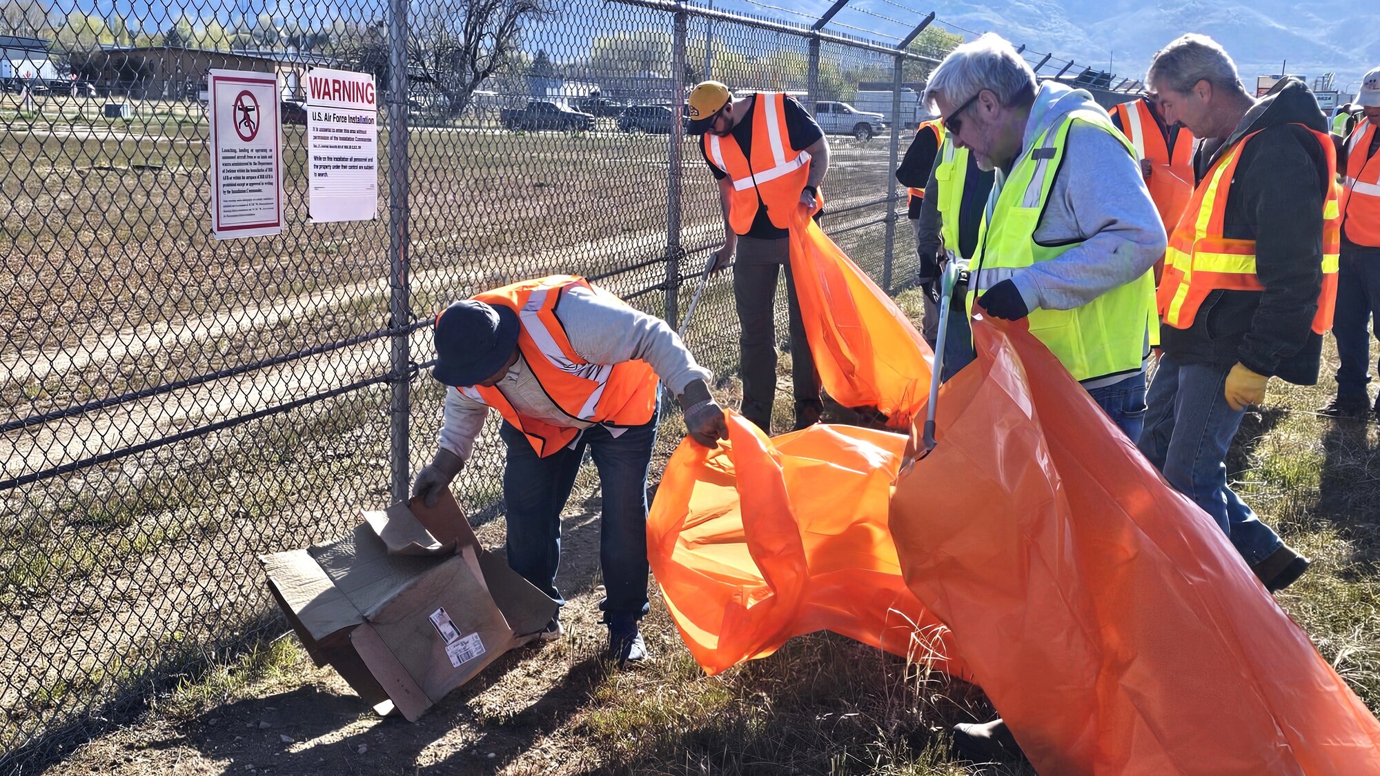 Volunteers, some holding trash bags, pick up trash along a fence line.