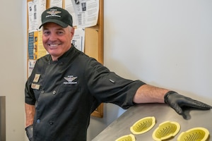 An Air National Guard chef poses for a photo in the Jefferson Barracks ANGS dining facility. He is wearing a black chef's coat and hat, standing next to a rack for organizing cutlery.