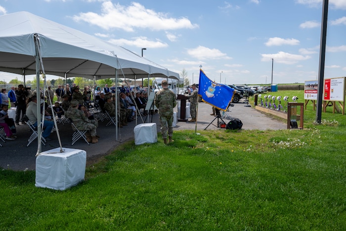 A man in a military uniform stands at a podium to deliver a speech to a group of people who are under an awning.