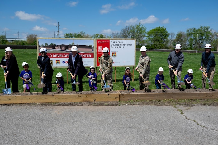 13 people stand in a row with shovels to symbolize the groundbreaking of a new piece of constriction.