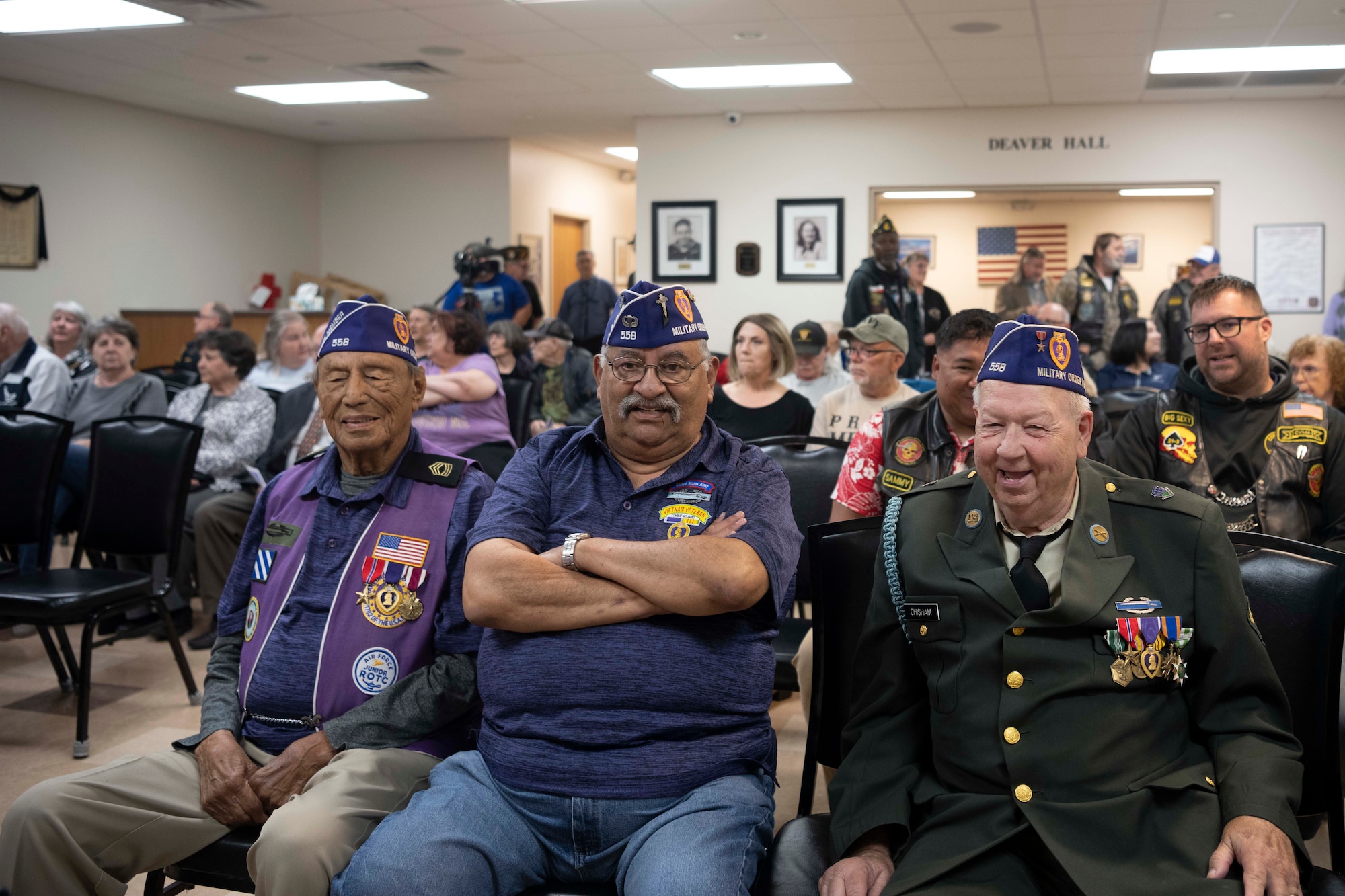 A group of Veterans are sitting in a conference hall wearing their purple vests signifying their purple heart status.