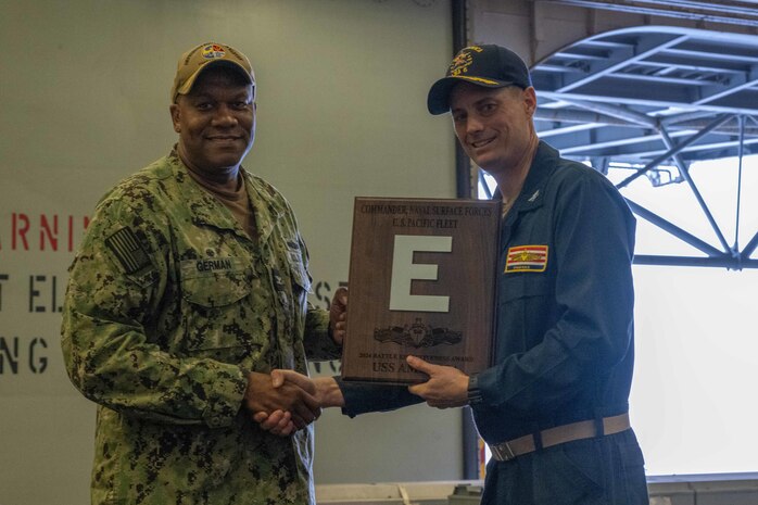 Capt. Ethan M. Rule, commanding officer of USS America (LHA 6), right, poses for a photo alongside Capt. Patrick German, Commodore Amphibious Squadron 11 (CPR 11), left, with the battle efficiency award for the forward-deployed amphibious assault ship USS America (LHA 6), in the ship’s hangar bay, April 14.