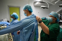 U.S. Air Force Senior Master Sgt. Nicole Nagle, 433rd Aeromedical Staging Squadron superintendent of health services assists Samuel Hobbs, 59th Medical Wing vitreoretinal surgeon, with putting on a sterile surgical gown before ophthalmology surgery at the Georgetown Public Hospital, Georgetown, Guyana, April 7, 2025 during the Lesser Antilles Medical Assistance Team 2025 mission.