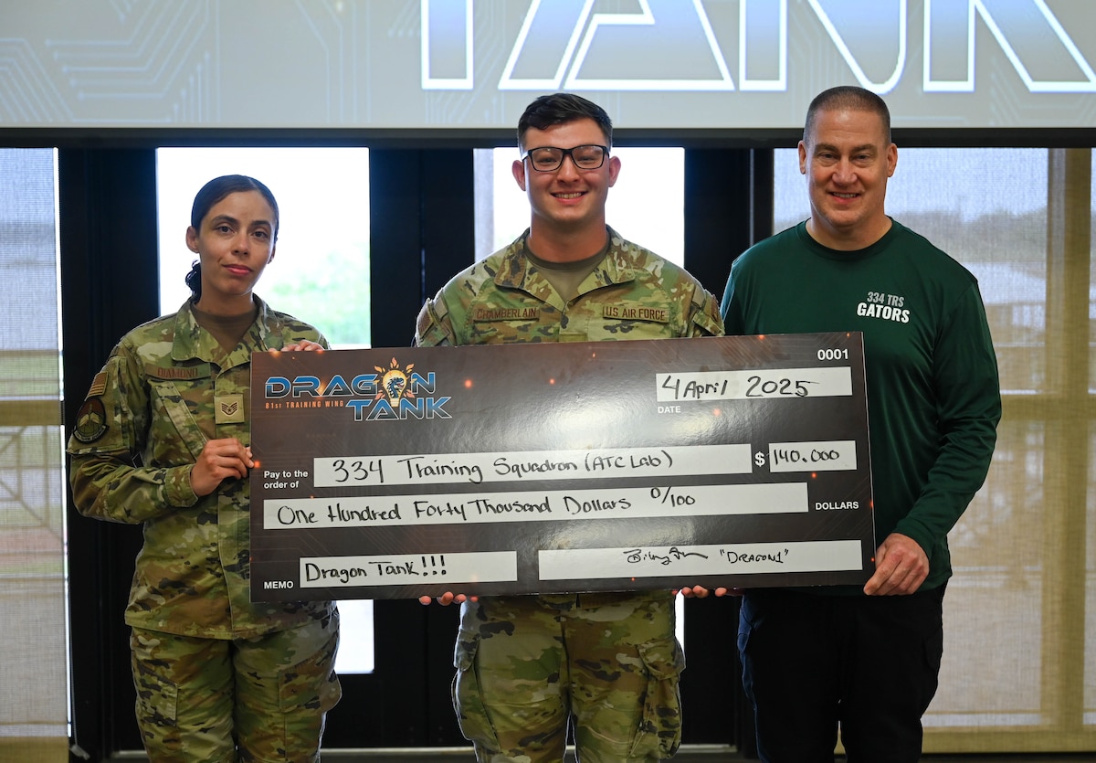 Two uniformed Airmen and one civilian pose with a large check for $140,000 after winning the Dragon Tank competition.