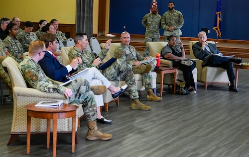 Six people sit in chairs during the Dragon Tank competition panel, with a uniformed male member, holds a microphone while speaking to presenters.