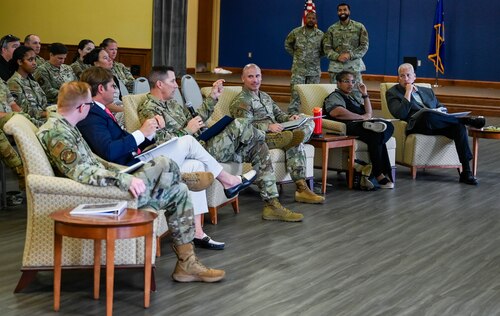 Six people sit in chairs during the Dragon Tank competition panel, with a uniformed male member, holds a microphone while speaking to presenters.