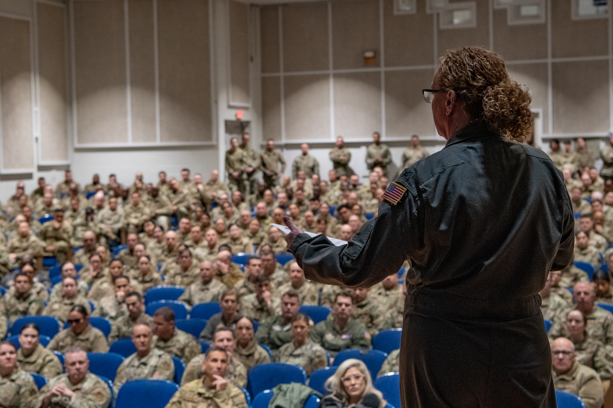 A woman in a flight suit speaking to a crowd of military personnel in theater seating.